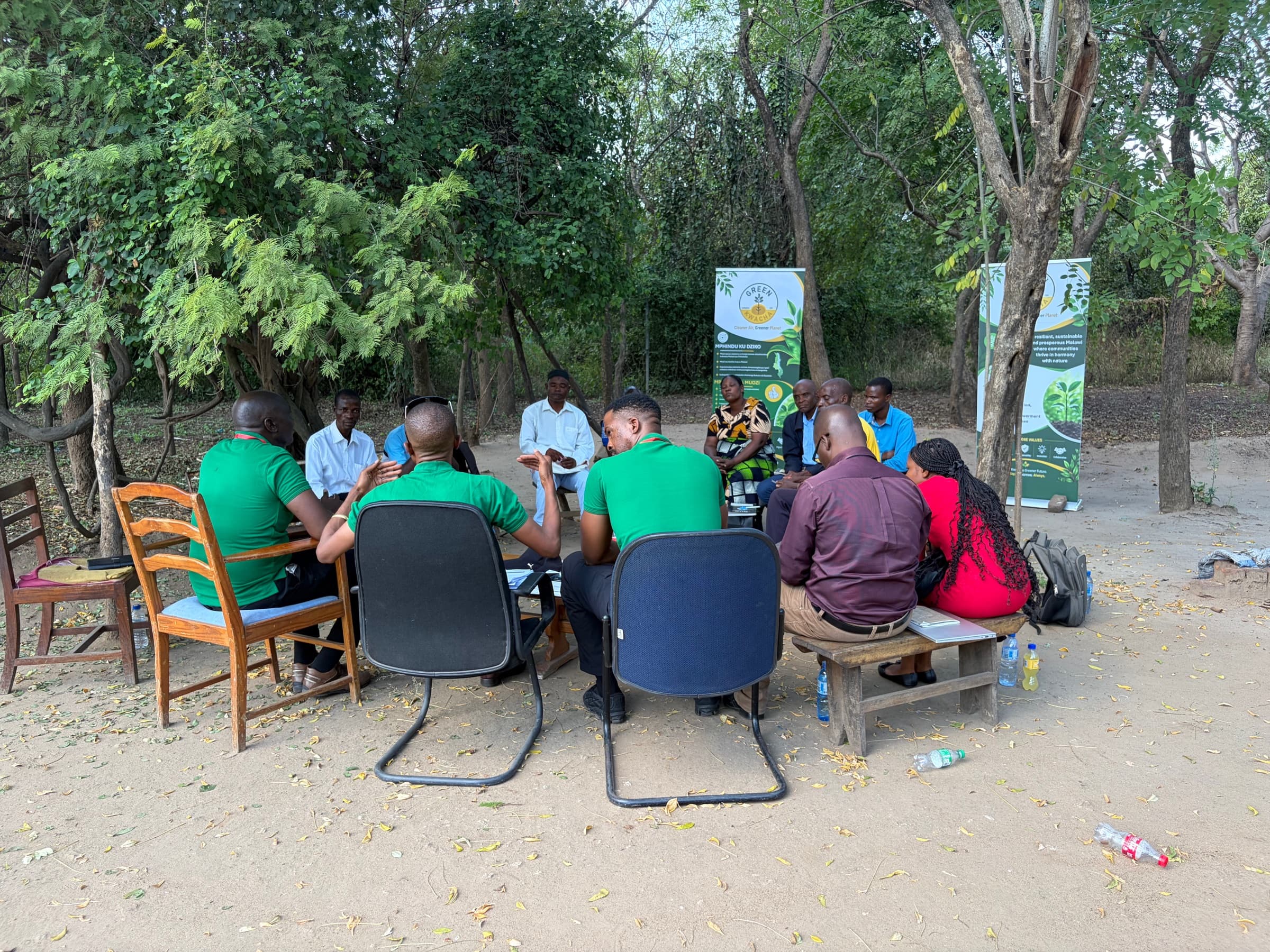 A wide view of a GreenKwacha listening circle with community members seated under a large tree, branded banner at the edge.